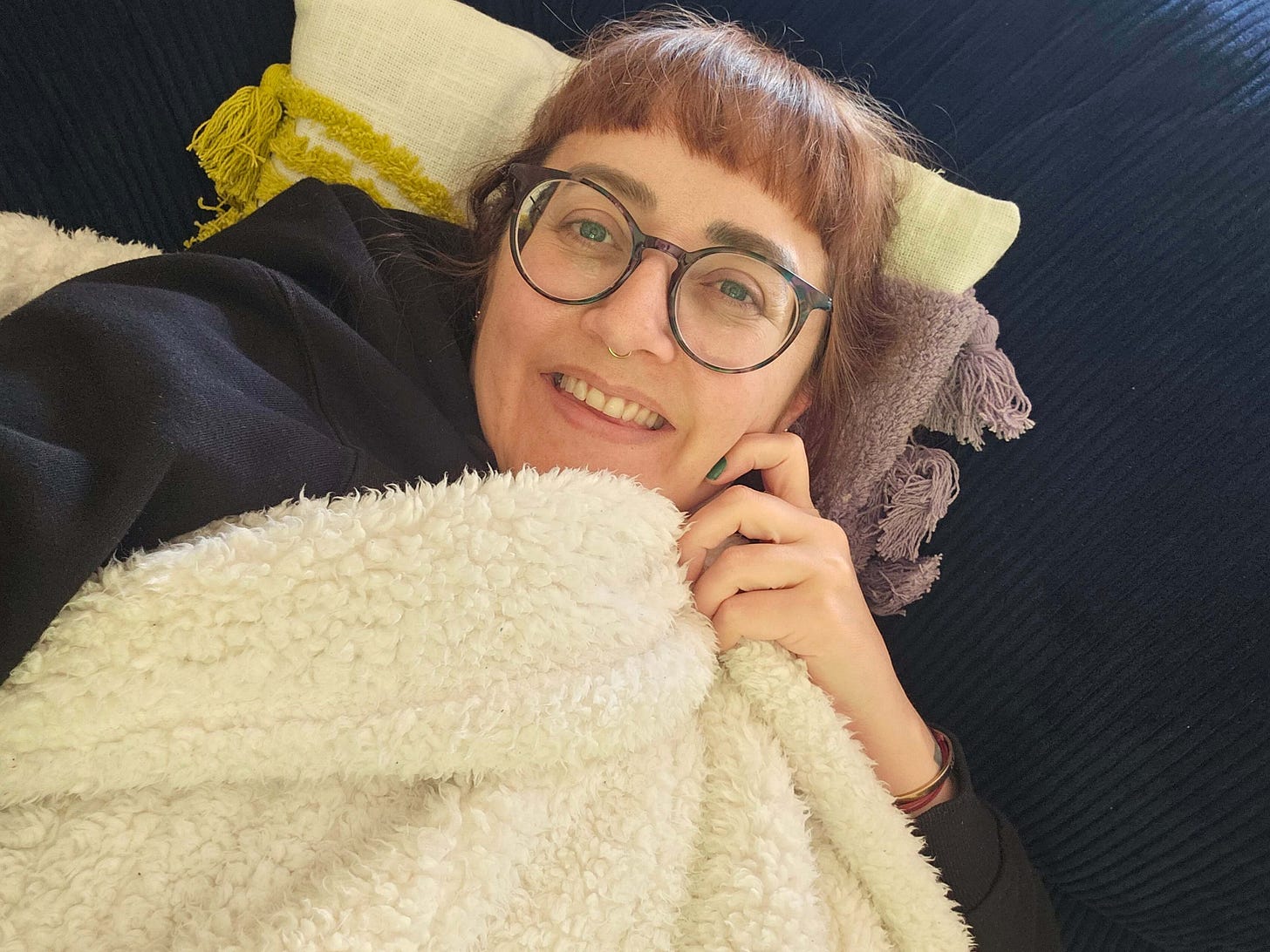 Sophie lying on a blue sofa with a white blanket pulled up to her chin. She is wearing glasses and smiling. Sophie lying on a blue sofa with a white blanket pulled up to her chin. She is wearing glasses and smiling.