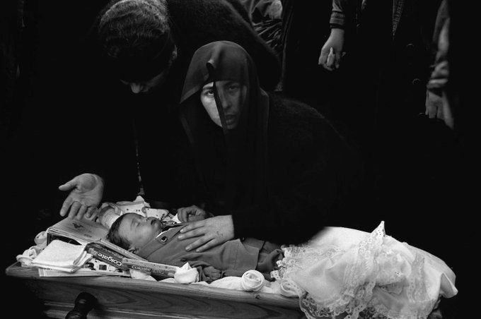 Black-and-white image shows a small wooden coffin on a table containing an infant wrapped in white lacy cloth with face visible eyes closed and hands crossed over chest surrounded by scattered papers and fabrics on the table surface several adults including men in hooded dark garments and a woman in black veil lean over the coffin with hands extended palms up in gesture of mourning and farewell additional figures stand in background wearing traditional clothing