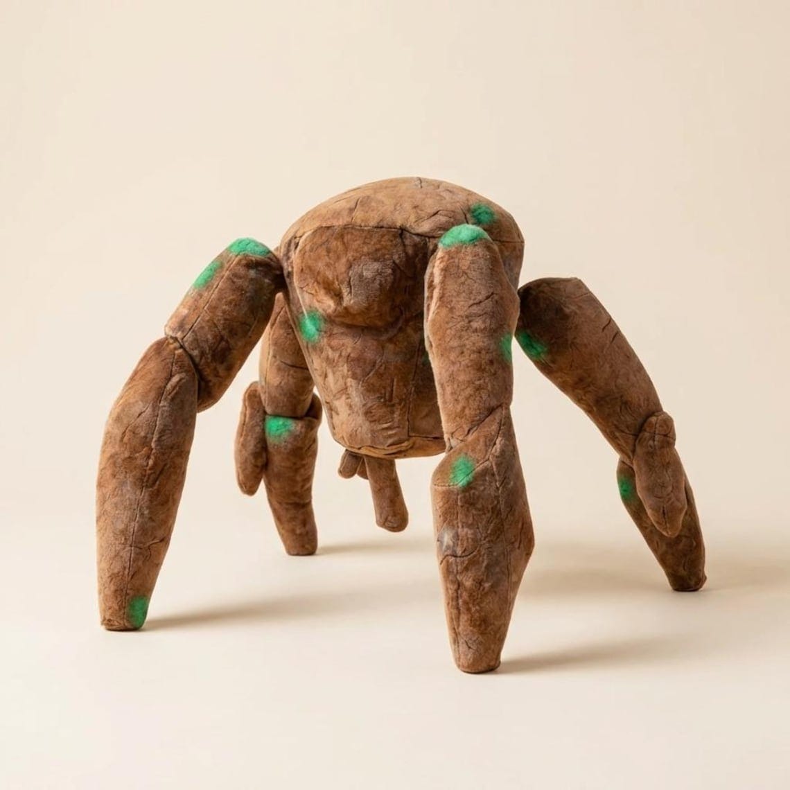 May include: A large, plush brown spider-shaped chair with six legs. The chair has green accents on the joints and body. The soft, textured material gives the chair a unique, whimsical appearance. The chair is designed for seating.