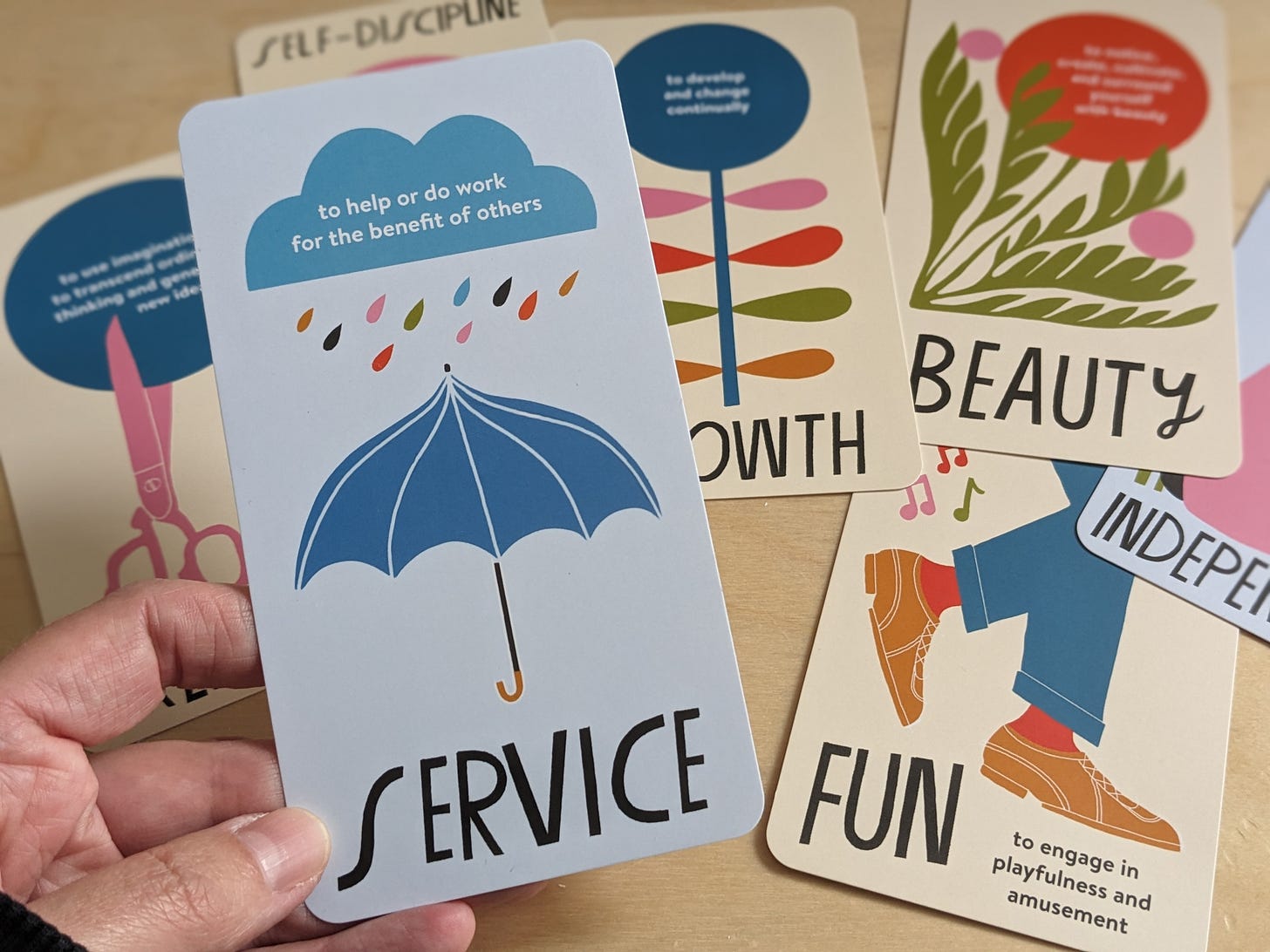 Image shows cards from Lisa Congdon's Live Your Values Card deck. The foremost card reads, Service. To help or do work for the benefit of others. The illustration on this card shows a raincloud raindrops coming out of it, with an umbrella underneath. Other cards are in the background, and read growth, fun, self-discipline, independence. They spill out of the sides of the photograph.