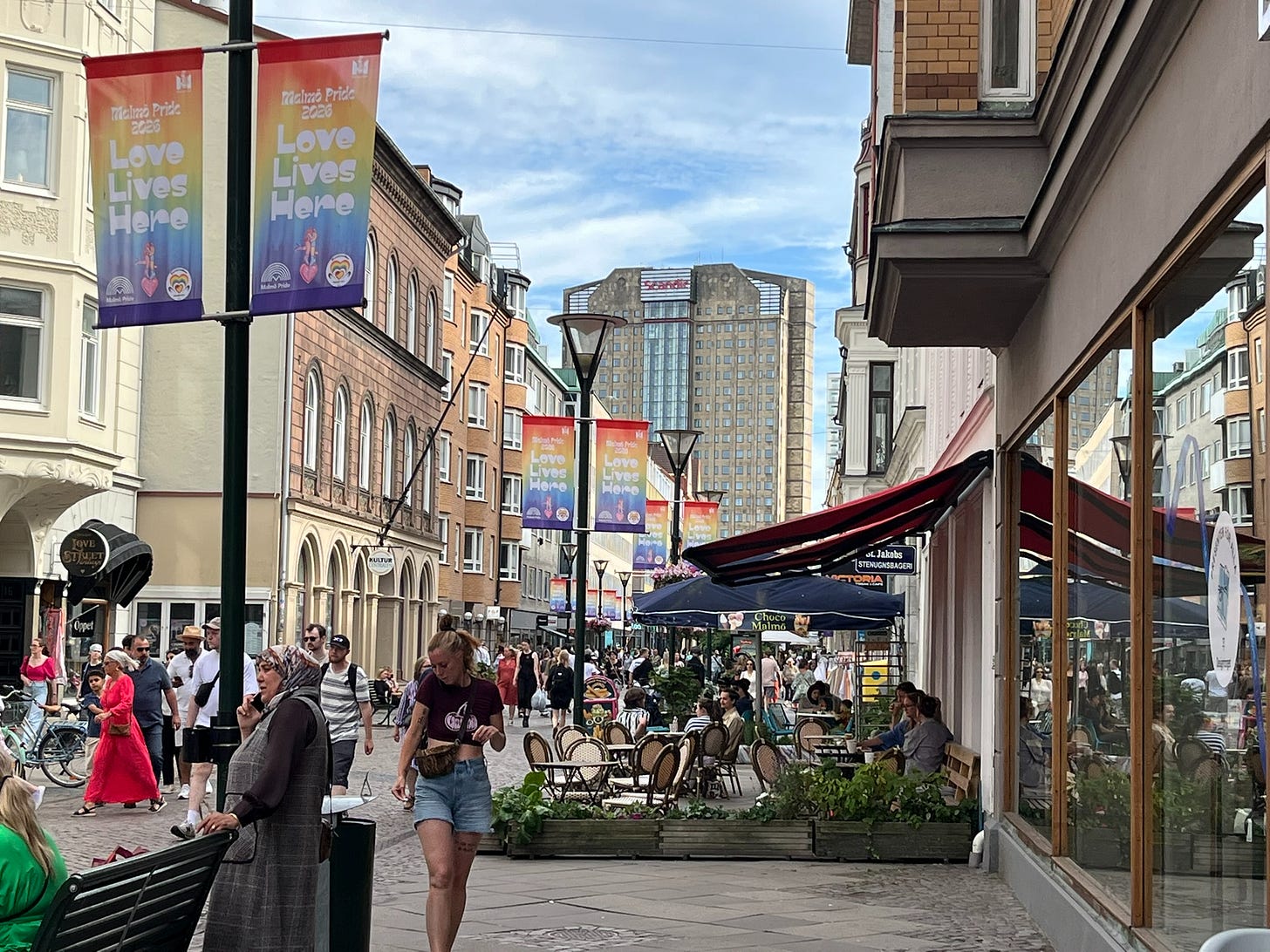 A largely pedestrian street with sidewalk cafes, with old mid-rise buildings on both sides, and a large gray concrete tower in the background with a red sign for the Scandic hotel chain 