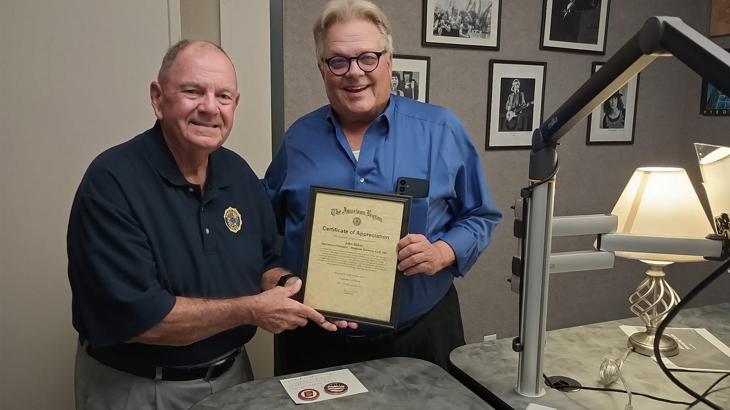 Two men are standing together indoors, smiling at the camera. The man on the left is wearing a dark polo shirt with a badge logo, while the man on the right is wearing glasses and a blue button-up shirt. They are jointly holding a framed "Certificate of Appreciation" from The American Legion, awarded to John Baker. The setting appears to be an office or studio, with a microphone arm, a table, a lamp, and black-and-white photos of musicians on the wall in the background. Two men are standing together indoors, smiling at the camera. The man on the left is wearing a dark polo shirt with a badge logo, while the man on the right is wearing glasses and a blue button-up shirt. They are jointly holding a framed "Certificate of Appreciation" from The American Legion, awarded to John Baker. The setting appears to be an office or studio, with a microphone arm, a table, a lamp, and black-and-white photos of musicians on the wall in the background.