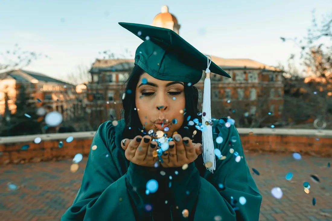 A joyful woman wearing a college graduation cap and gown and blowing sparkly confetti because she had just graduated.