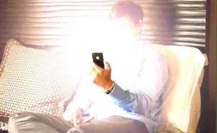 This may contain: a man sitting on a bed holding a cell phone in his hand and looking at the screen