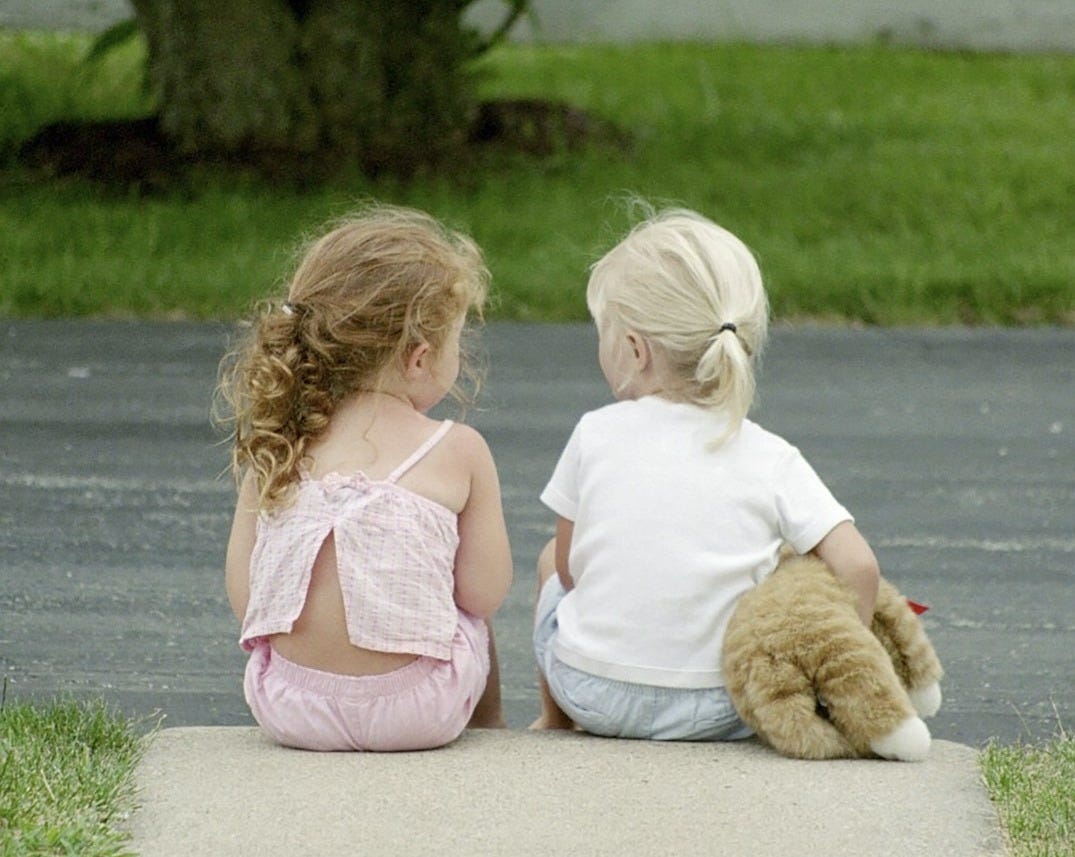 Two little girls sitting on the sidewalk with a teddy bear
