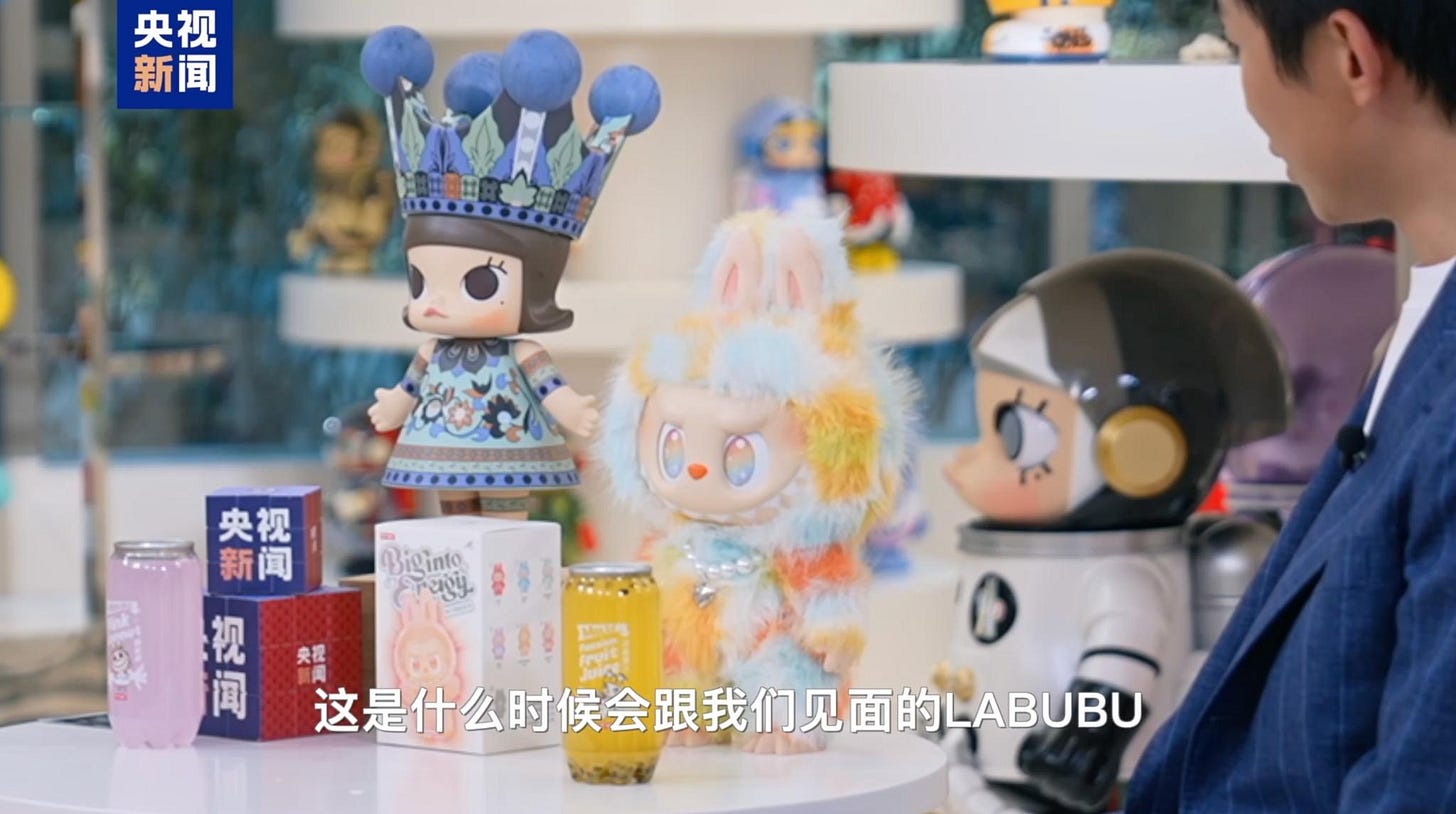 Close‑up of prototype Labubu plush in pastel faux‑fur next to other Pop Mart toys on a studio table; Chinese subtitle reads, “When will we meet this Labubu?” while a spokesperson in a blue blazer looks on. Close‑up of prototype Labubu plush in pastel faux‑fur next to other Pop Mart toys on a studio table; Chinese subtitle reads, “When will we meet this Labubu?” while a spokesperson in a blue blazer looks on.