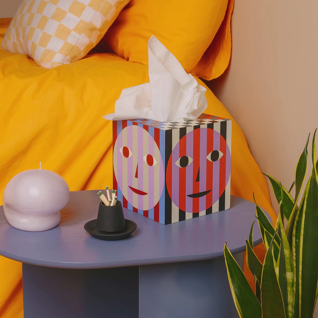 Dusen Dusen tissue box made up of various reds, blues, greens and beige lines and smiley faces. The tissue box is staged on a bedside table next to a blob candle and matches set up in front of a yellow bed Dusen Dusen tissue box made up of various reds, blues, greens and beige lines and smiley faces. The tissue box is staged on a bedside table next to a blob candle and matches set up in front of a yellow bed