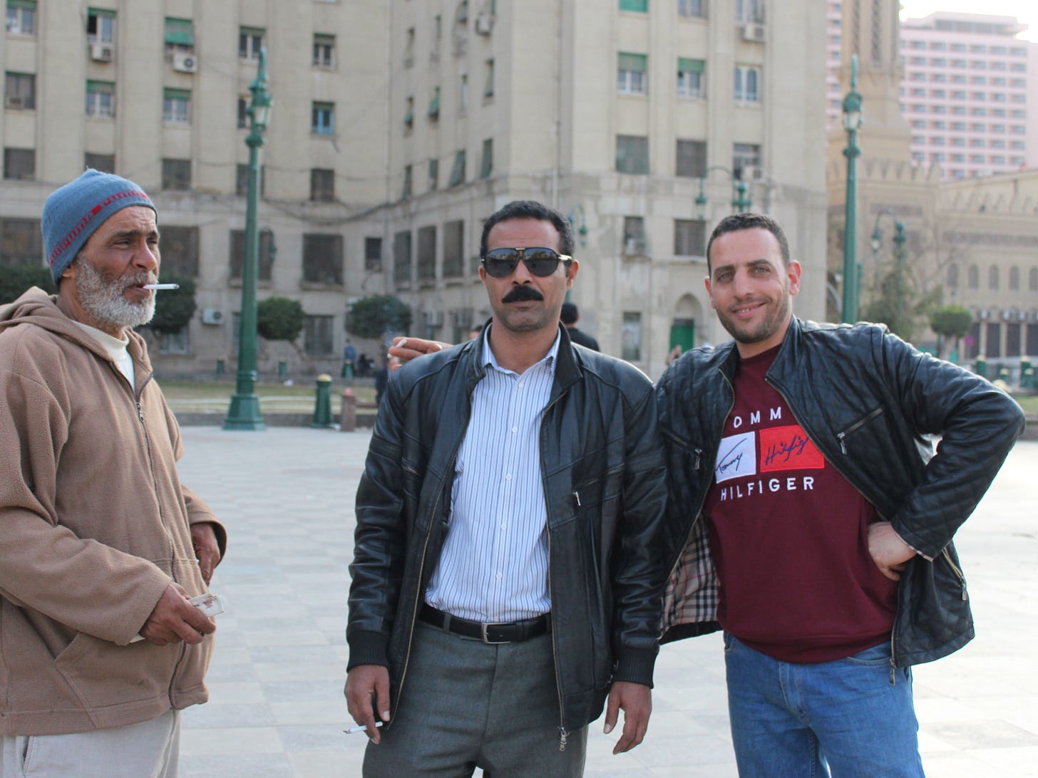 Three men smoke cigarettes and pose in Tahrir Square, Cairo, the one in the middle wearing aviator sunglasses and a black leather jacket, with the Mogamma government building behind them.