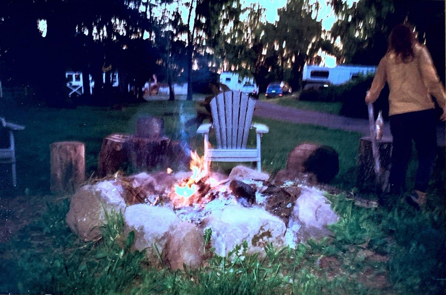 a 35 mm photo of a fire pit formed of big rocks with stumps around it for sitting and a couple of chairs. A person in a mustard yellow jacket and black pants is facing away from the camera holding a big fire poking stick. it's dusk.