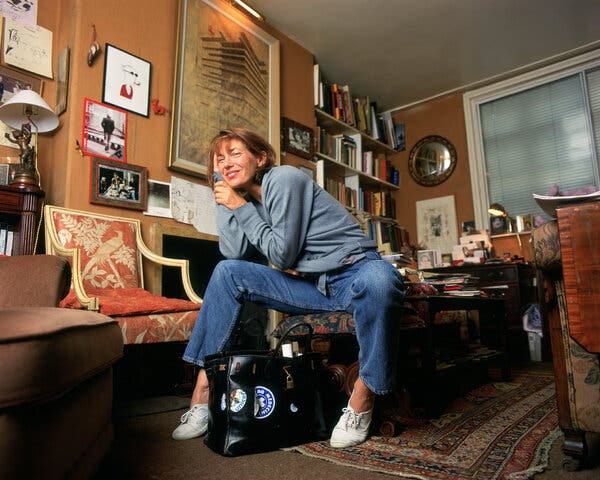 A woman in a blue sweatshirt, jeans and white shoes sits on a chair. Between her legs is a black bag covered in stickers. 