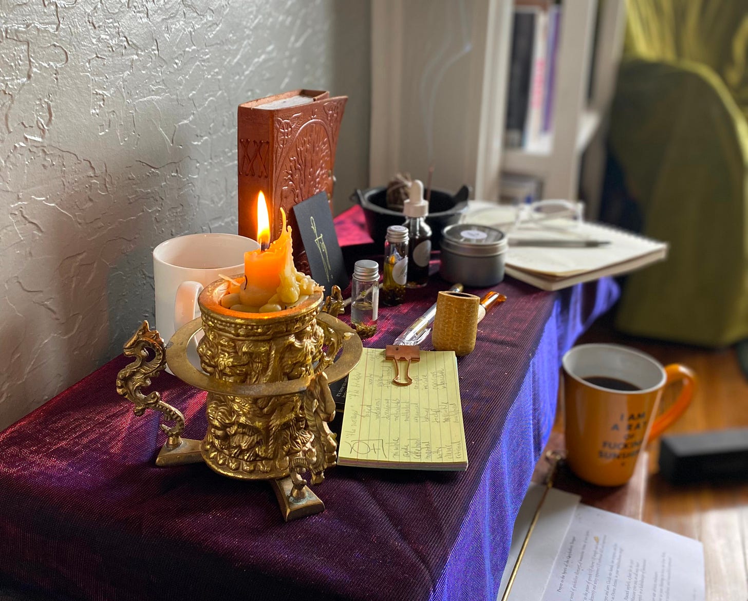a top down view of an altar covered in objects: small bottles of oil, a leatherbound journal, a sigil, a notebook, stack of notecards, coffee, a small cauldron with herbs burning inside, and a golden candle.
