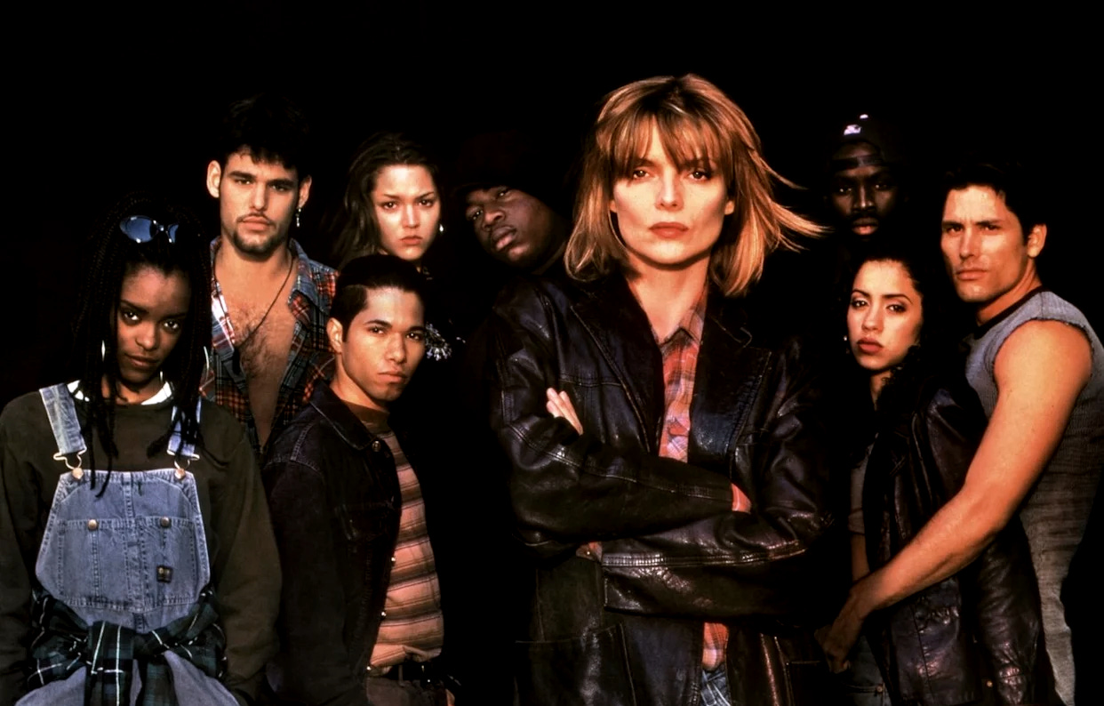 Cast photo from the 1995 film Dangerous Minds showing Michelle Pfeiffer in a leather jacket surrounded by eight student actors of diverse backgrounds, all looking seriously at the camera against a black background.