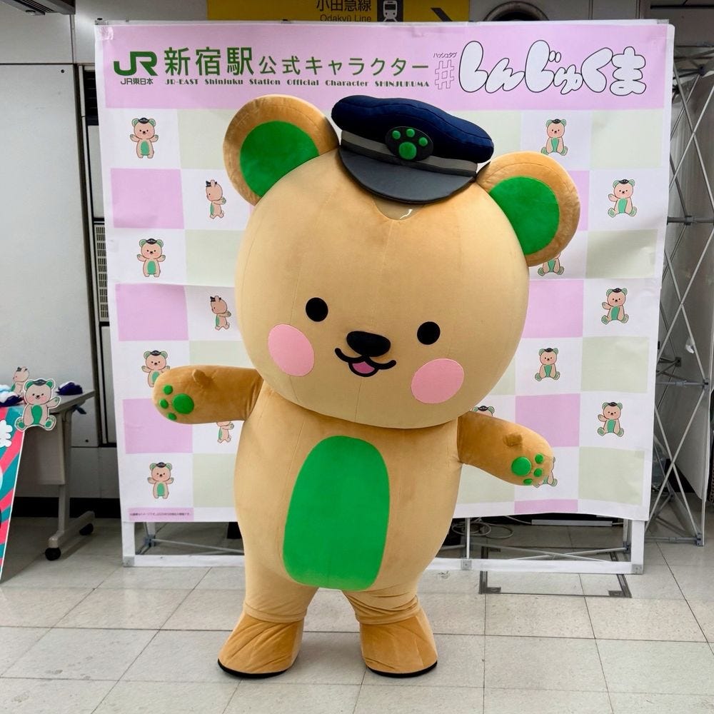 A beige bear mascot with green paws, belly, and ears, poses in a station master’s cap