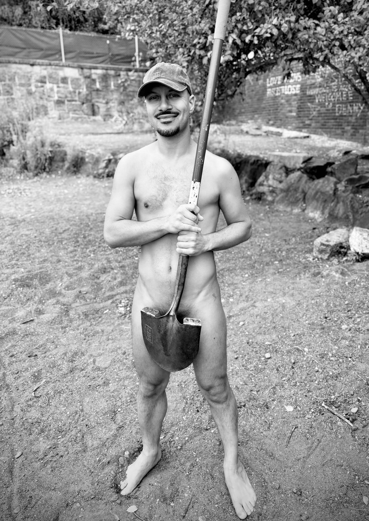 A nude person wearing a cap stands barefoot on a sandy beach, smiling and holding a large shovel upright in front of them. Behind them is a stone wall with fencing and trees, part of Denny Blaine Park in Seattle.
