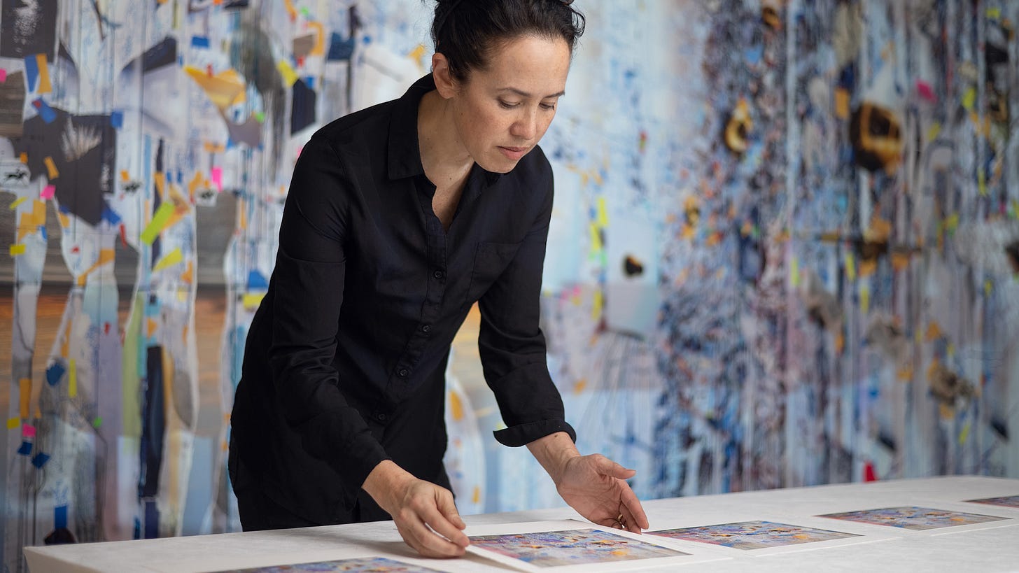 The artist Sarah Sze leans over a table, handling prints of her work. She has black hair pulled up, and is wearing all black. Behind her is a blurred image of one of her large, colorful, collage-style paintings