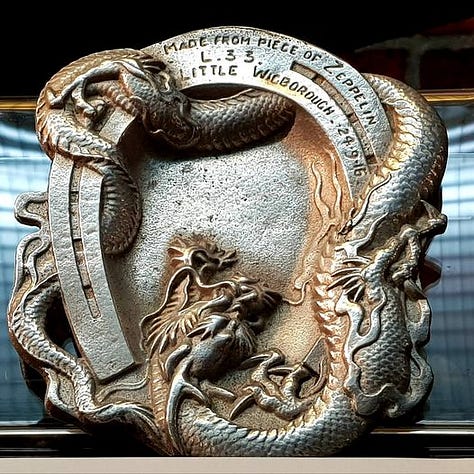 Image one shows the museum display for World War 1, featuring a model zeppelin. Image 2 shows an ornate metal ashtray. A dragon weaves in an out of a horse shoe which circles the entire edge. Top centre the inscription reads made from pieces of Zeppelin L33, Little Wigborough 24.9.16. The third image shows small pieces of the zeppelin's metal framing.