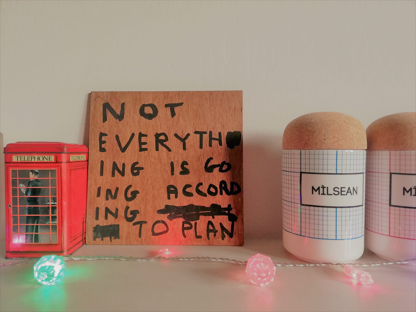 a mantlepiece with an old telephone box biscuit tin, a David Shrigley quote on a board - 'Not everything is going to plan' - and two storage containers