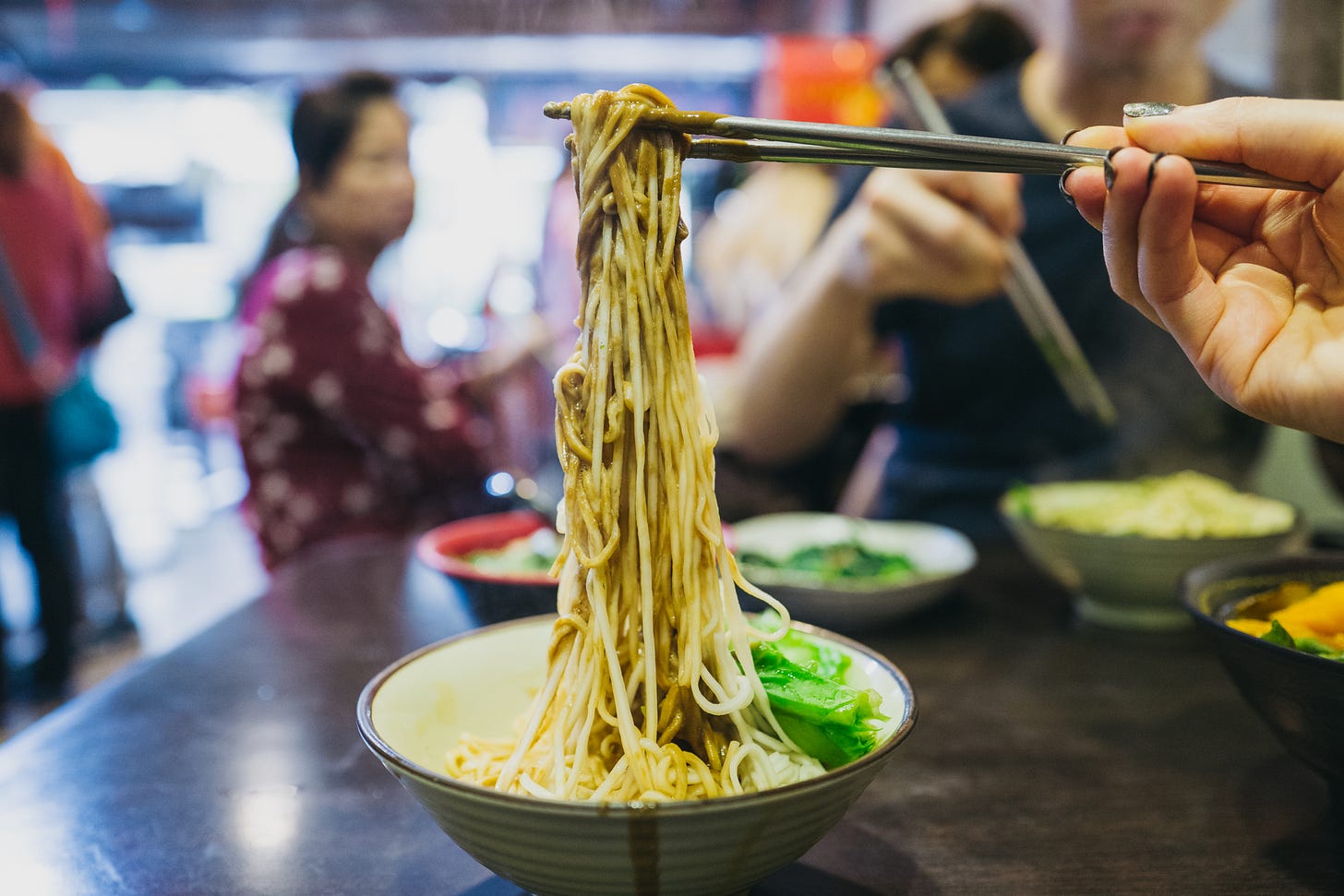 Chopsticks lifting a bundle of noodles from a bowl at a busy restaurant, with diners blurred in the background