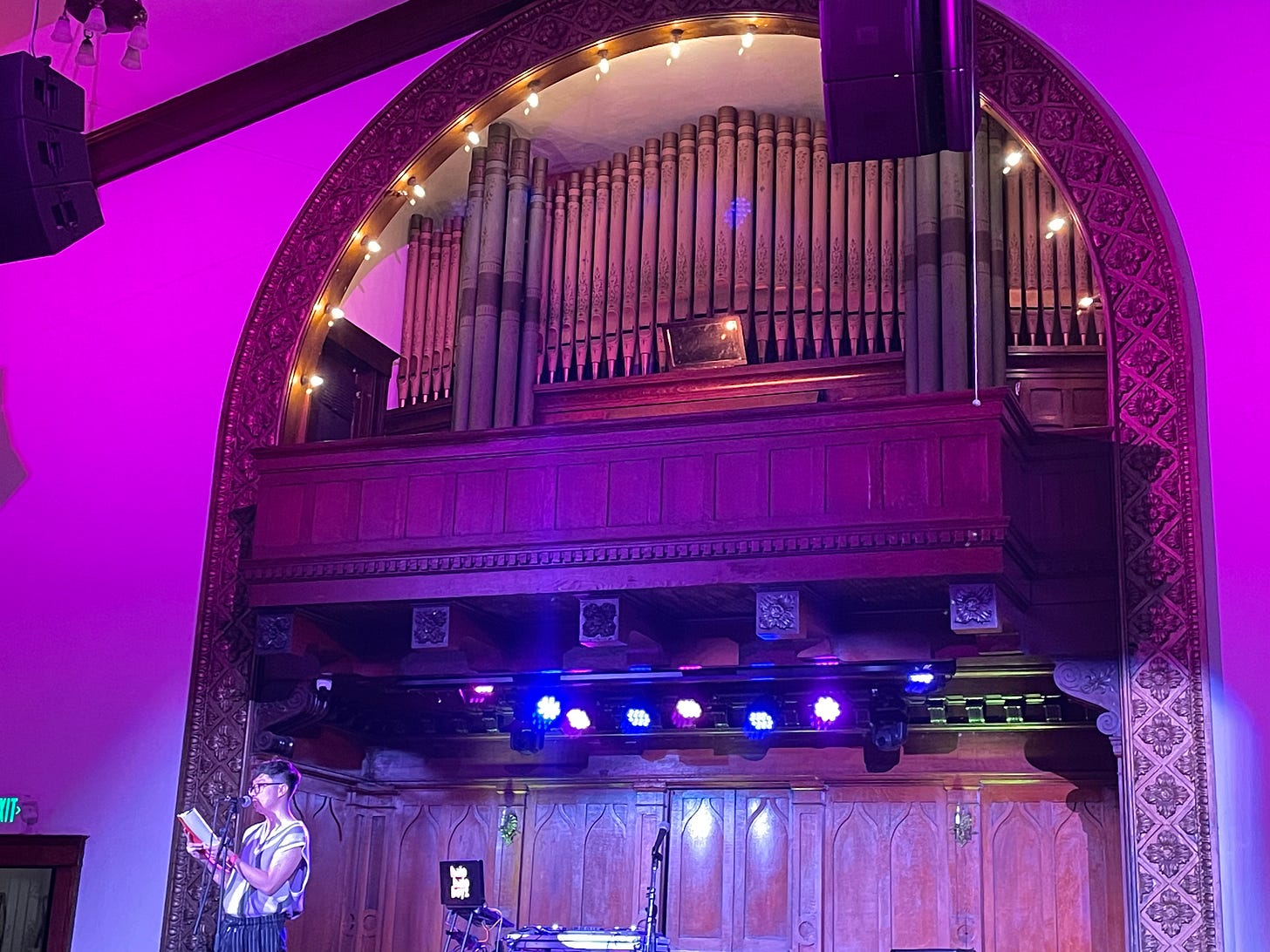 an Asian man poet in a patterned tank top reads in front of a church's pipe organ.