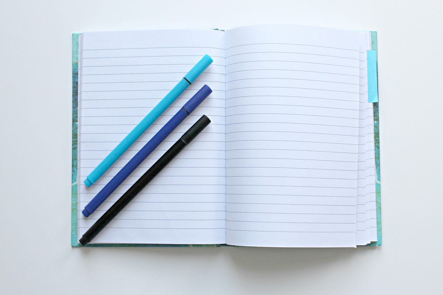 Three pens of different shades of blue laid diagonally on an open notepad with blank white pages
