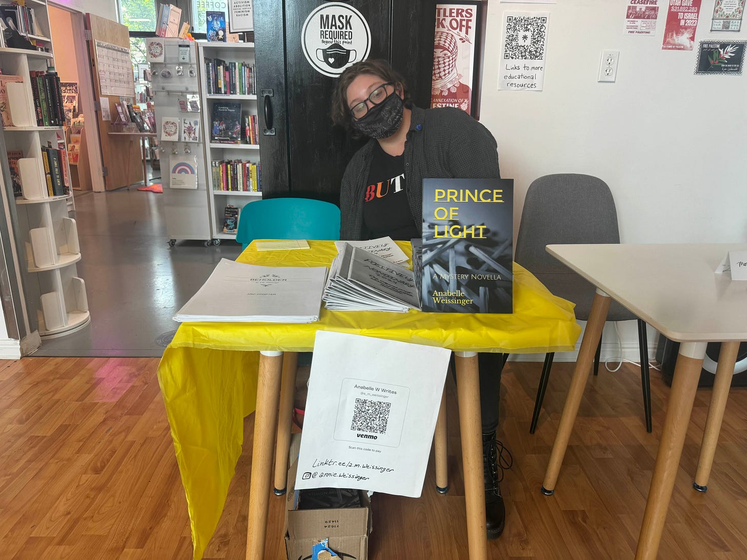 Mayson sits behind a table with a yellow tablecloth and stacks of books and zines.