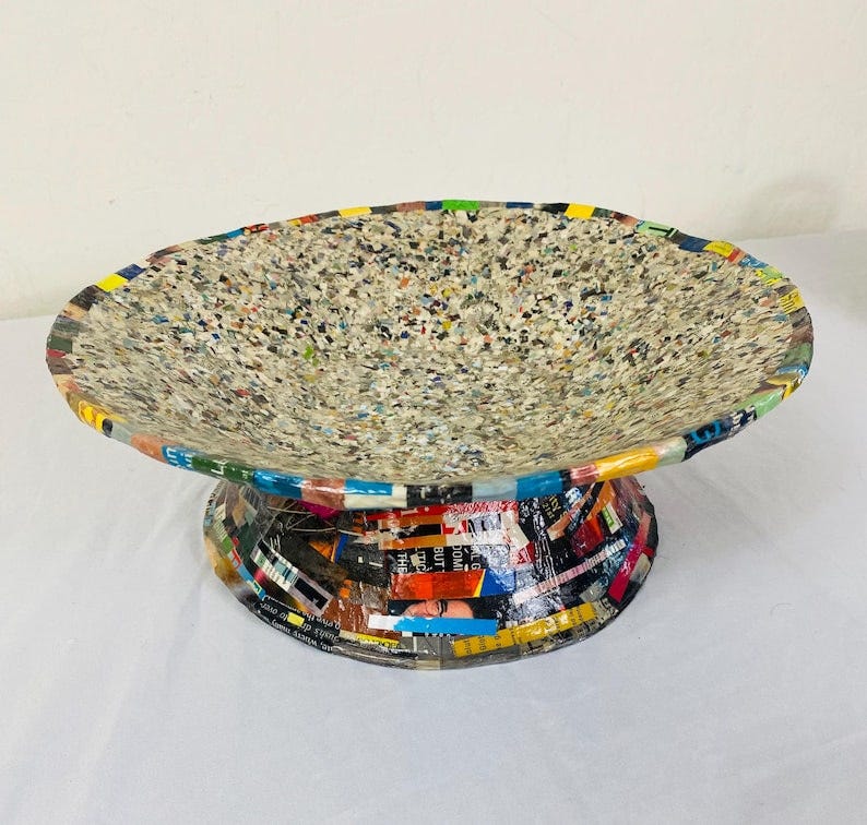 May include: A decorative pedestal bowl with a wide, shallow dish. The bowl's surface is speckled with a mix of colors, while the base is adorned with colorful paper strips. This unique piece is ideal for displaying fruit or other decorative items.