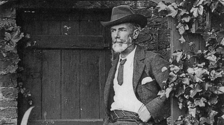 Black-and-white photograph of Edward Carpenter seated outdoors beside a wooden door framed with climbing ivy. He wears a wide-brimmed hat, jacket, tie, and vest, with one hand resting on his hip. He has a neatly trimmed beard and mustache and gazes off to the side with a thoughtful expression.