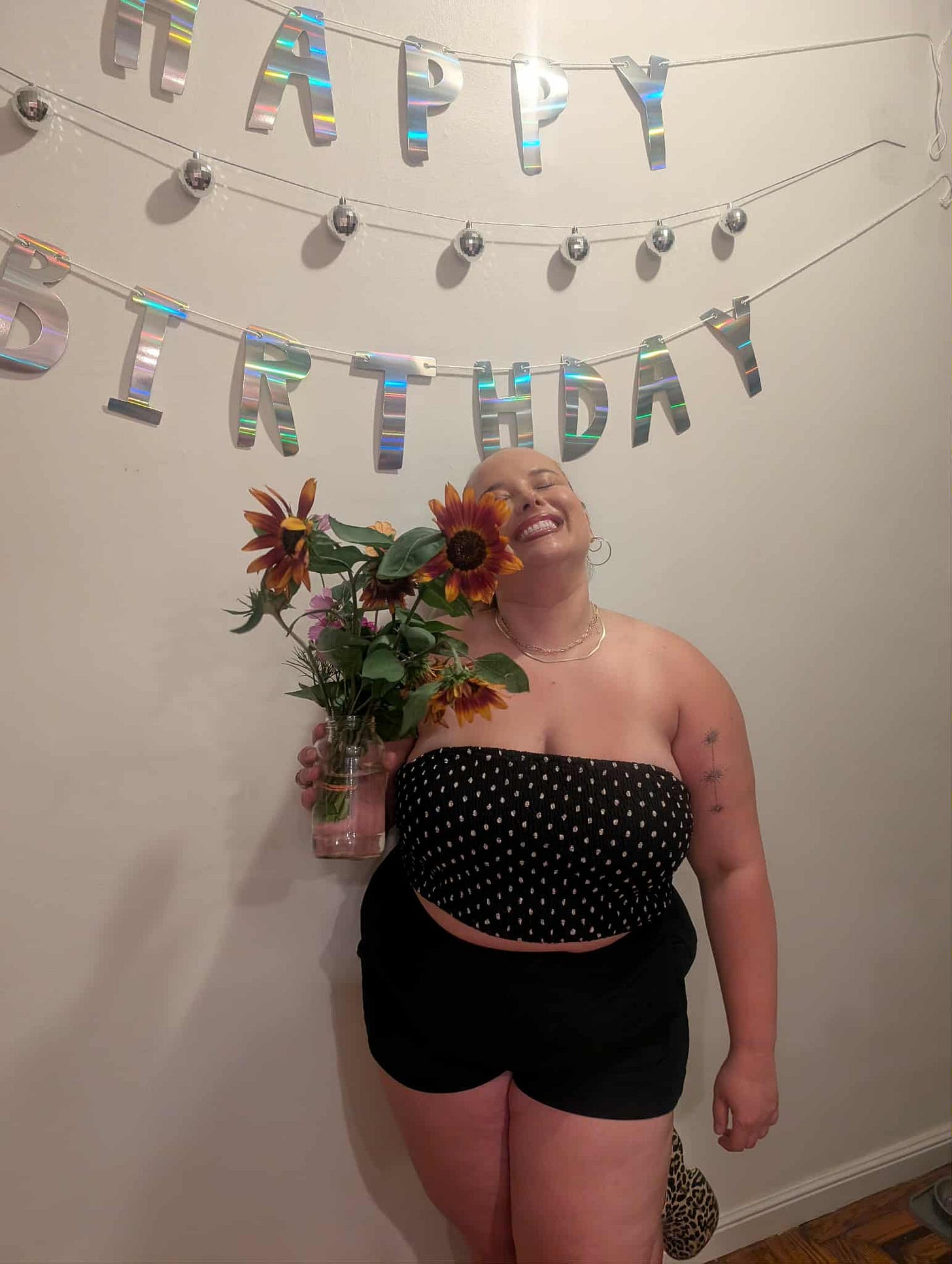 Kate (a white person with a polka dot strapless top and black shorts)is smiling brightly while holding a bouquet of sunflowers. They’re standing in front of a wall decorated with shiny “HAPPY BIRTHDAY” letters and string lights.