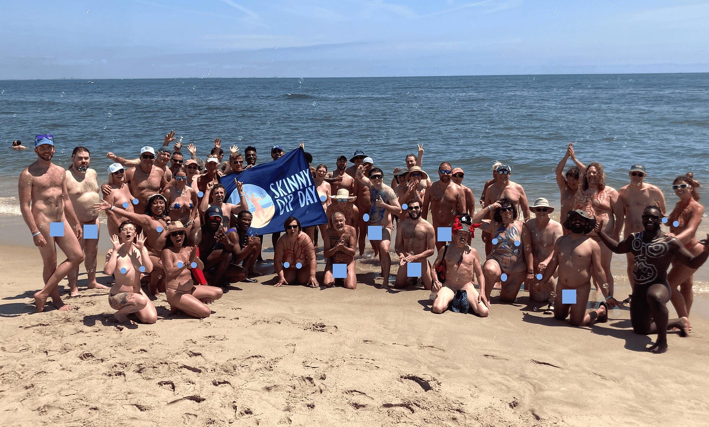 A large group of nude participants pose together on the sand at Gunnison Beach, New Jersey, holding a Skinny Dip Day banner. The sky is blue and the ocean calm behind them. Many people are smiling, raising their arms, or crouching in front, celebrating their group skinny dip. A large group of nude participants pose together on the sand at Gunnison Beach, New Jersey, holding a Skinny Dip Day banner. The sky is blue and the ocean calm behind them. Many people are smiling, raising their arms, or crouching in front, celebrating their group skinny dip.