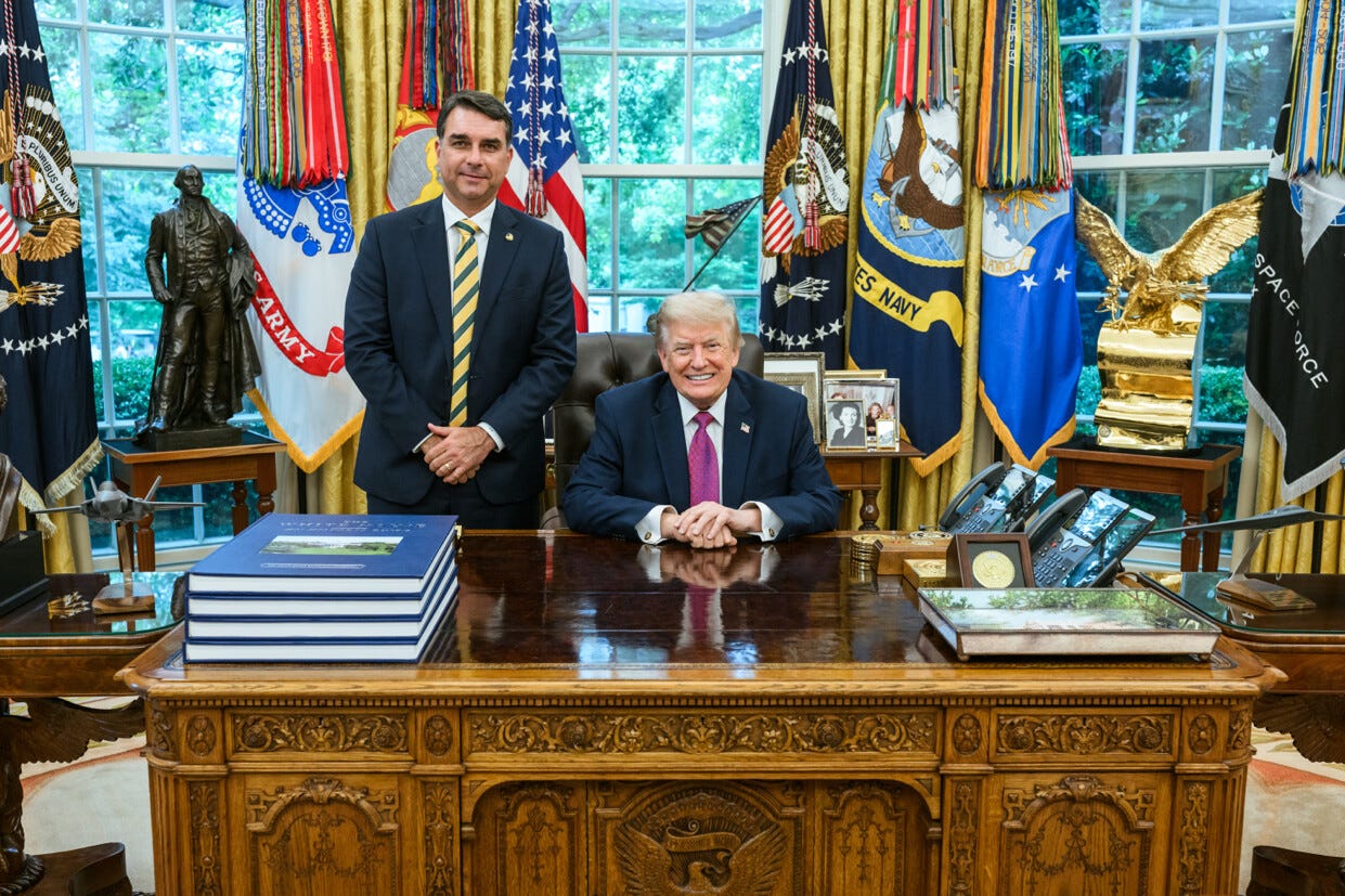 Flávio Bolsonaro stands beside Donald Trump, who is seated behind the Resolute Desk in the Oval Office.