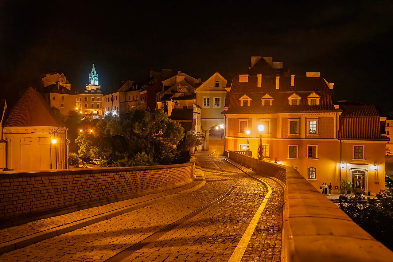 Photo: Krzysztof Popławski / Wikimedia Commons (CC BY-SA 4.0) Night view of the Old Town and city centre of Lublin , Lublin Voivodeship , Poland , on 23 August 2025. The photograph shows a cobblestoned street running through the historic buildings of the Old Town, a retaining wall, tenement houses, illuminated