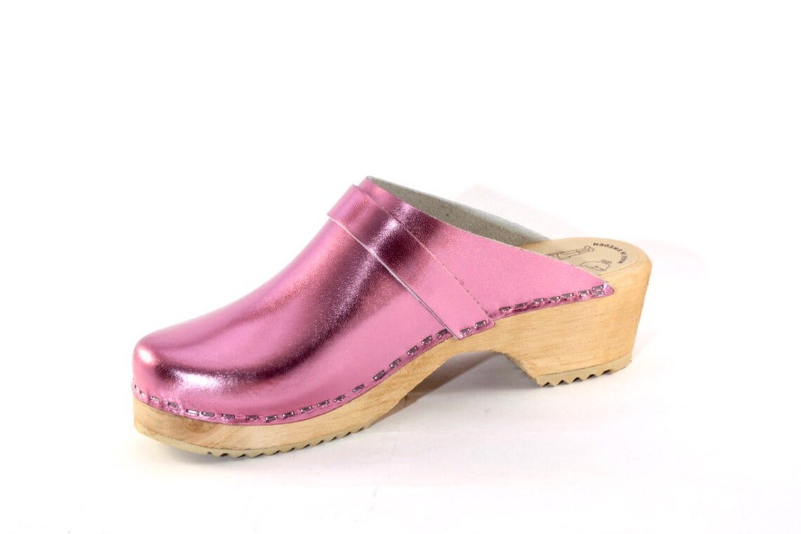May include: Pink metallic clog with a wooden sole. The shoe features a rounded toe and a strap across the top. The wooden sole has a slight heel and a textured bottom. May include: Pink metallic clog with a wooden sole. The shoe features a rounded toe and a strap across the top. The wooden sole has a slight heel and a textured bottom.