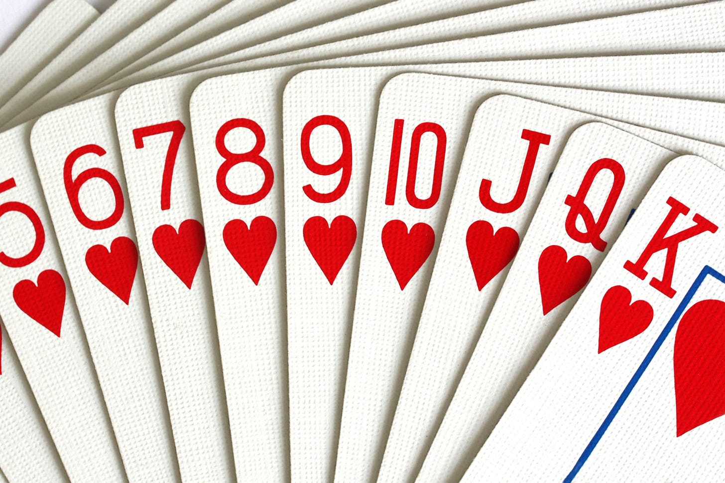 A close-up, fan-shaped arrangement of playing cards from the suit of Hearts. The cards visible range from the Five of Hearts to the King of Hearts, shown in sequential order from left to right. The cards have a textured, linen-finish surface, and the red Heart symbols and numbers are bold and vibrant against the white background. A close-up, fan-shaped arrangement of playing cards from the suit of Hearts. The cards visible range from the Five of Hearts to the King of Hearts, shown in sequential order from left to right. The cards have a textured, linen-finish surface, and the red Heart symbols and numbers are bold and vibrant against the white background.