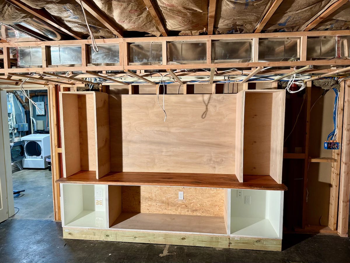 An unfinished built-in entertainment center shows the bottom cabinetry in place along with an oak countertop, while the upper cabinetry is temporarily placed.