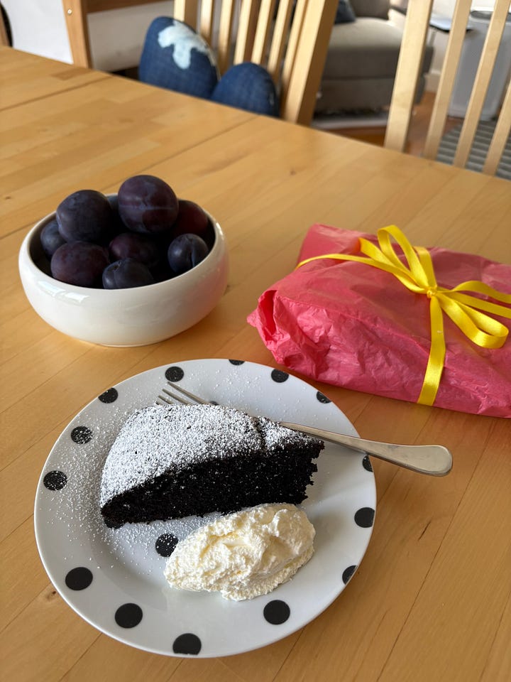 Four plates of yoghurt cake with strawberries and a slice of chocolate olive oil cake alongside a bowl of plums and a present
