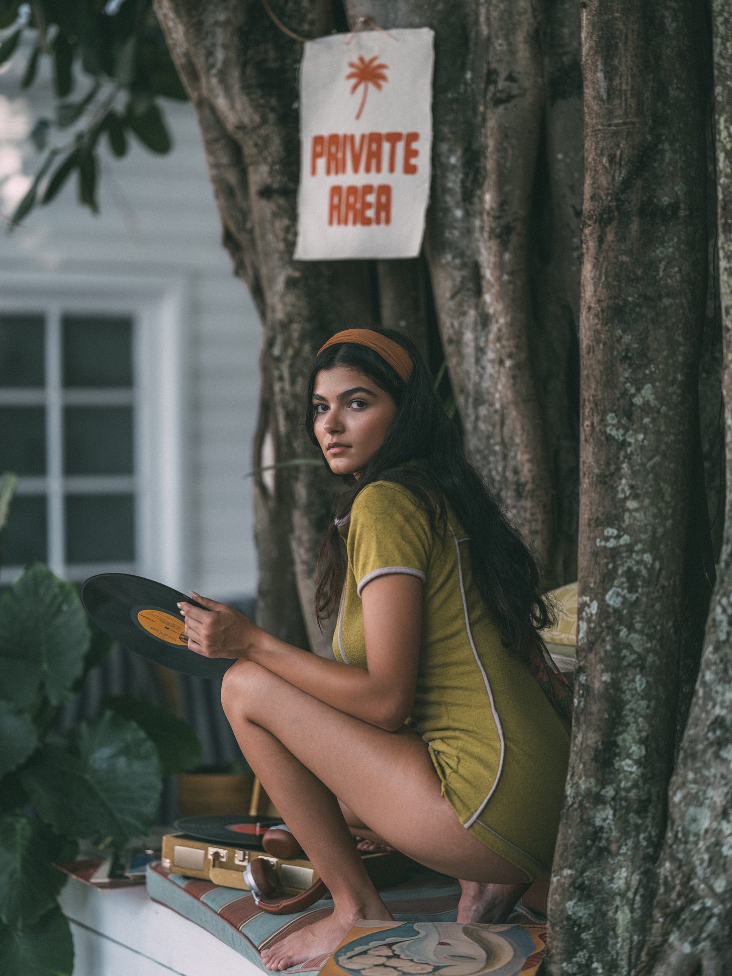 A woman with dark hair crouches next to a large tree while holding a vinyl record. She wears a green vintage-style romper and an orange headband, and looks at the camera. A sign on the tree above her reads "PRIVATE AREA."