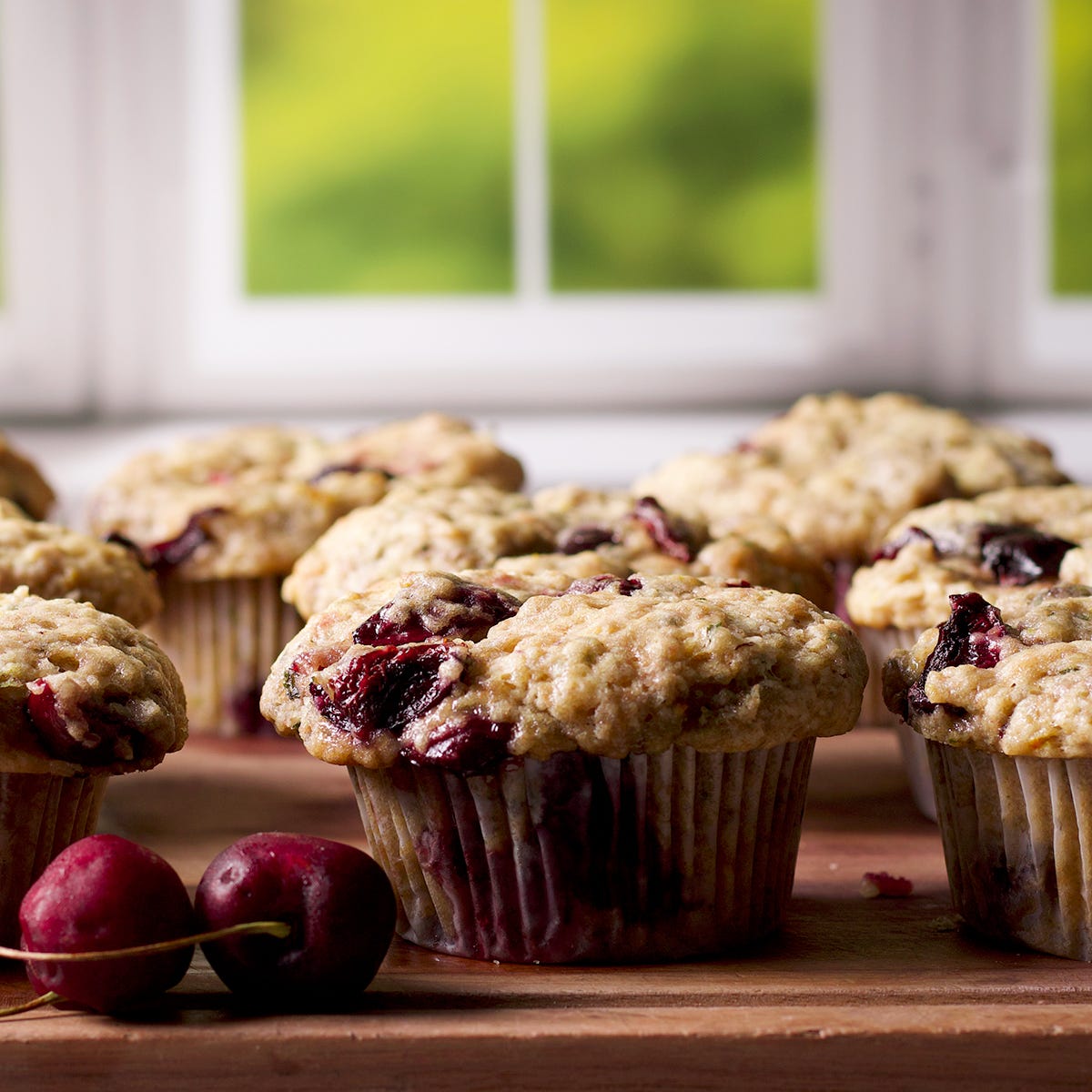 Homemade cherry muffins cooling on a wooden board next to a window.