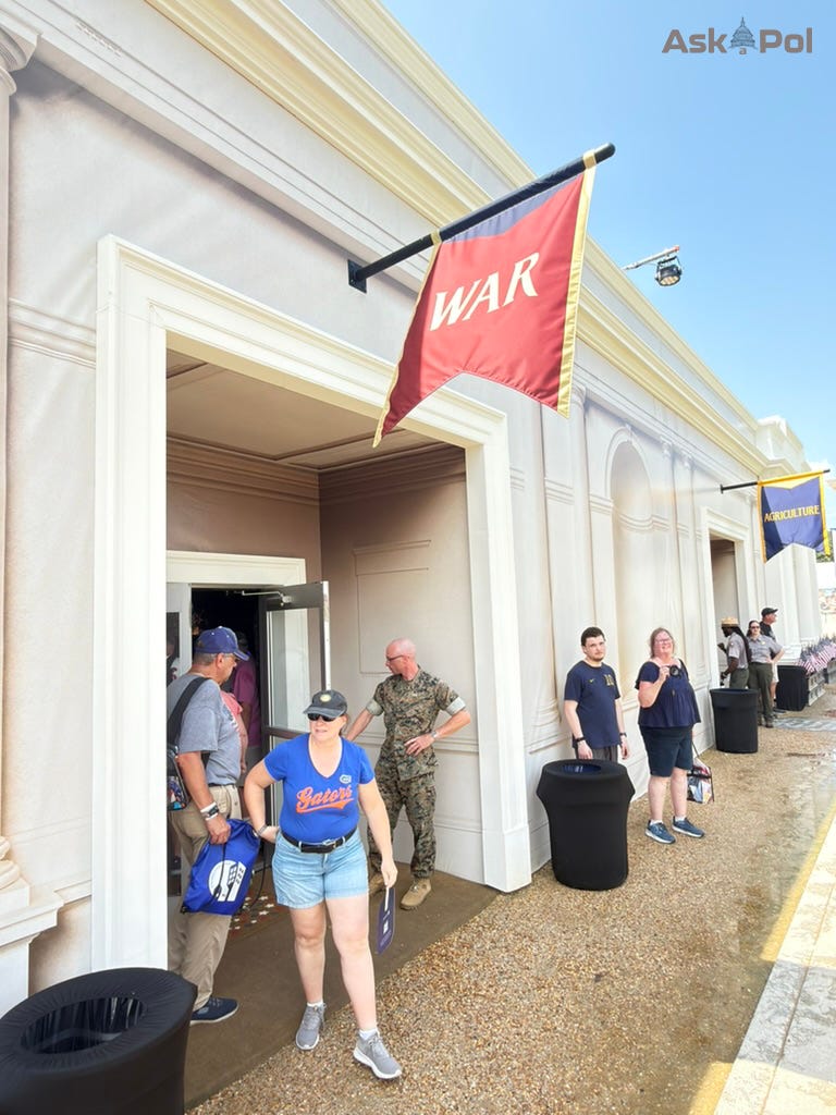 A red "WAR" flag laps in the wind on a sunny day, as visitors huddle in the shade and a soldier holds the door to the exhibit open at Freedom 250 in DC A red "WAR" flag laps in the wind on a sunny day, as visitors huddle in the shade and a soldier holds the door to the exhibit open at Freedom 250 in DC