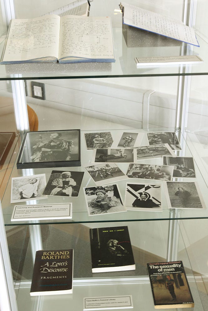 Glass display case with three shelves. The top shelf holds open notebooks with handwritten notes. The middle shelf showcases black-and-white photographs of individuals. The bottom shelf displays three books, titled "A Lover’s Discourse," "The Sexuality of Men," and one with an unreadable title. Each shelf has a text placard giving more context about the items on the shelf.