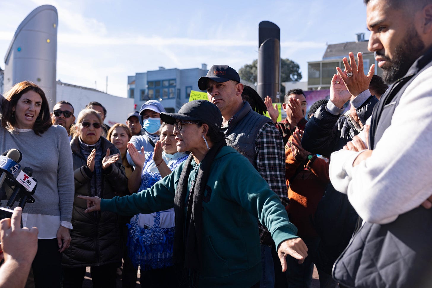 A woman named Gladys points in both directions at waist level with a large group of people listening to her, including Supervisor Hillary Rosen to her right.