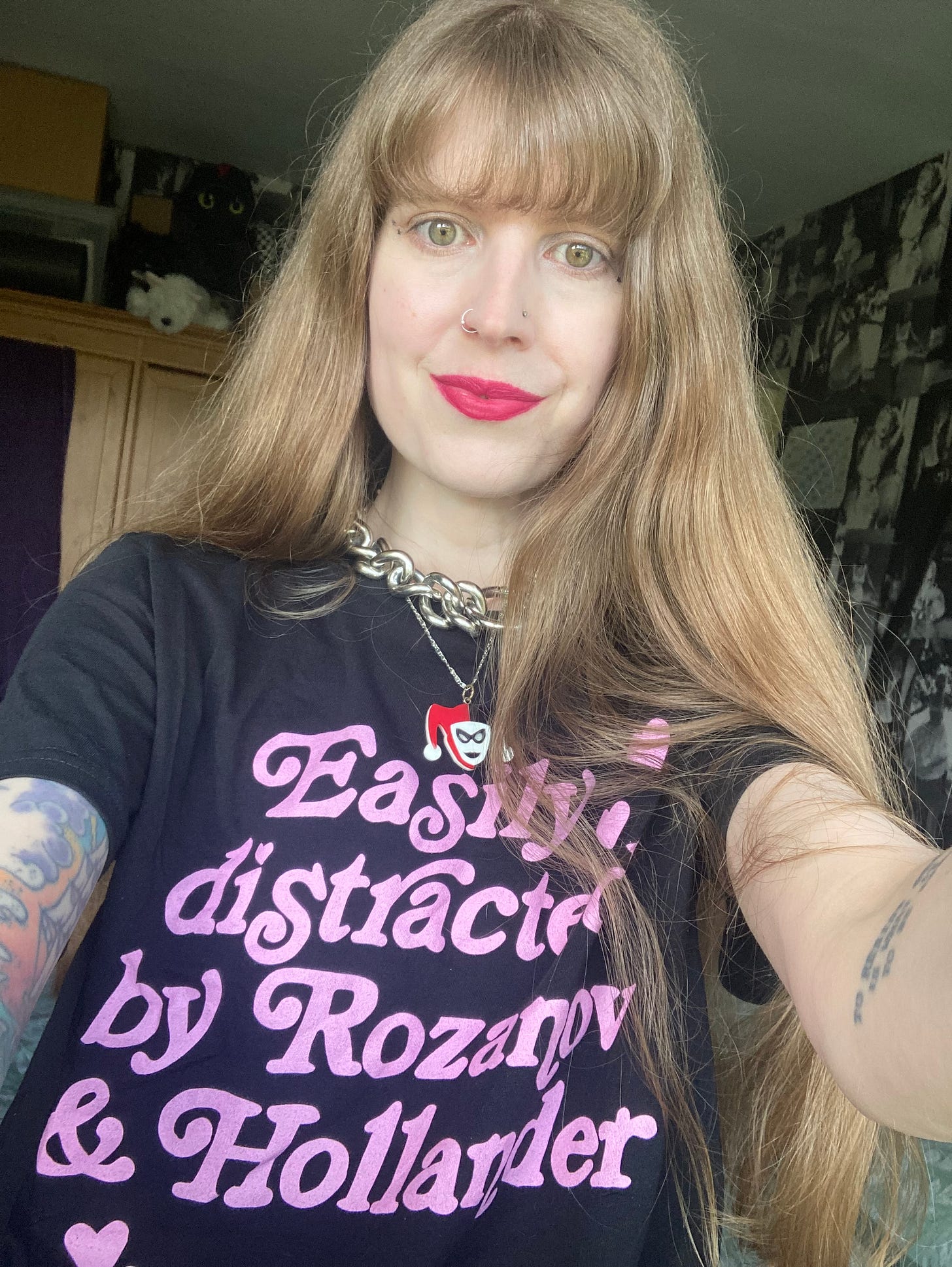 The writer looks out from the photo. They have long blonde hair and are wearing pink lipstick, a silver chain necklace and a Harley Quinn necklace. They are wearing a black t-shirt with pink writing. The writing says "Easily distracted by Rozanov and Hollander" who are characters from Heated Rivalry. The writer looks out from the photo. They have long blonde hair and are wearing pink lipstick, a silver chain necklace and a Harley Quinn necklace. They are wearing a black t-shirt with pink writing. The writing says "Easily distracted by Rozanov and Hollander" who are characters from Heated Rivalry.