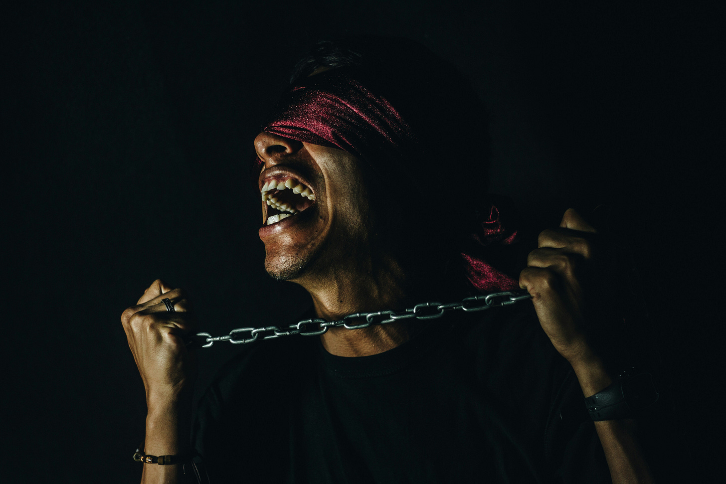 A person wearing a red blindfold grips a metal chain across their neck while laughing, symbolizing the paradoxical nature of being trapped in hustle culture. The dramatic lighting and dark background emphasize the emotional struggle of breaking free from self-imposed work constraints.