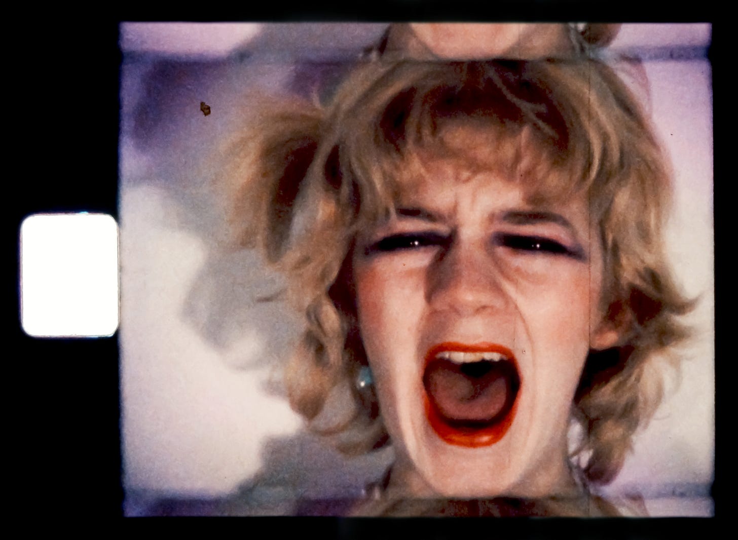 A still of a white blonde woman screaming in anger and anguish on a film camera reel. Her red lipstick is smudged on her teeth.  Image credit: Gina Birch, still from 3 Minute Scream, 1977. Courtesy of the artist. Image courtesy of: National Galleries Scotland