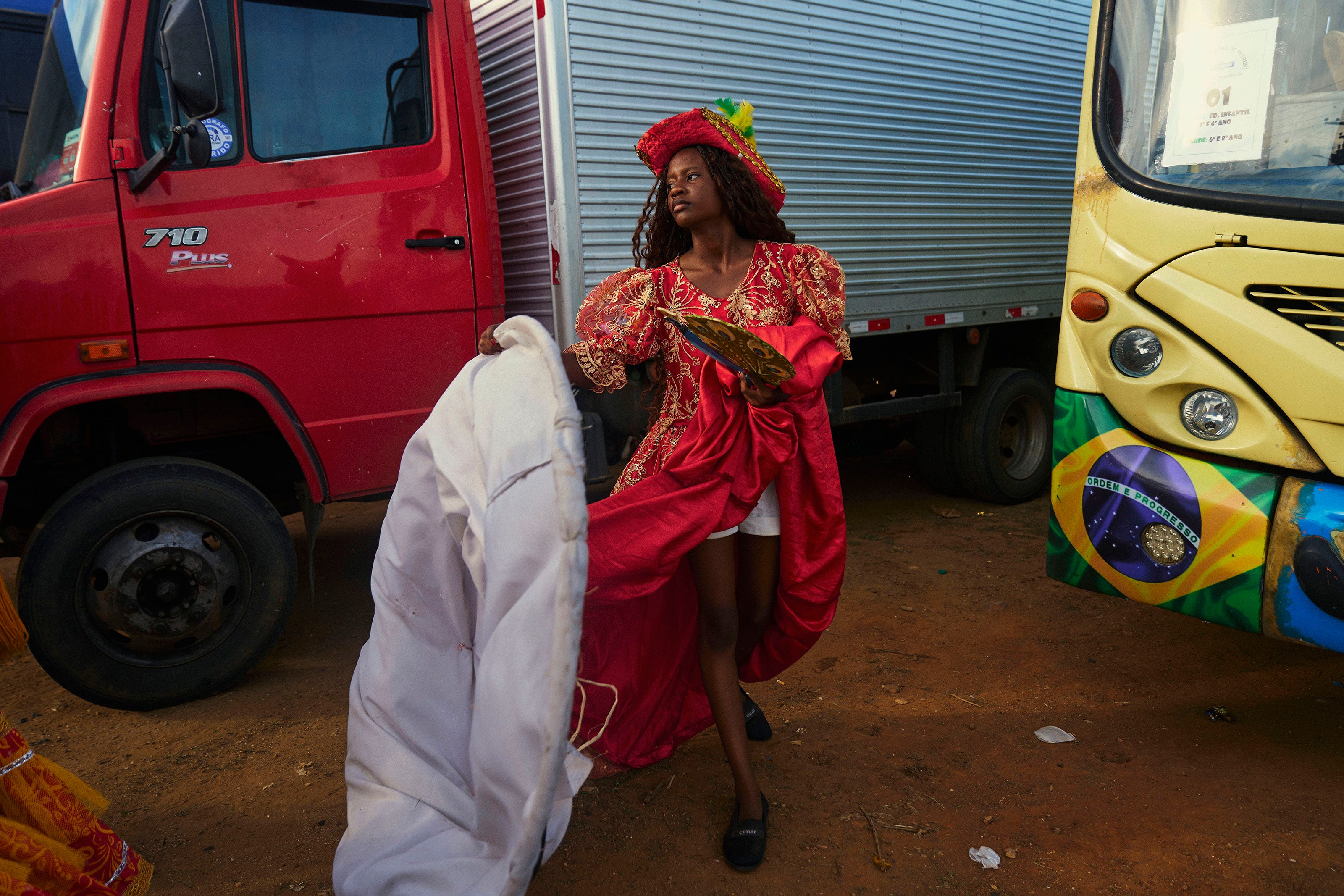 A woman in a vibrant red costume with gold details and a matching hat stands between a red truck and a yellow bus decorated with a Brazilian flag, holding a white cloth and looking to the side.