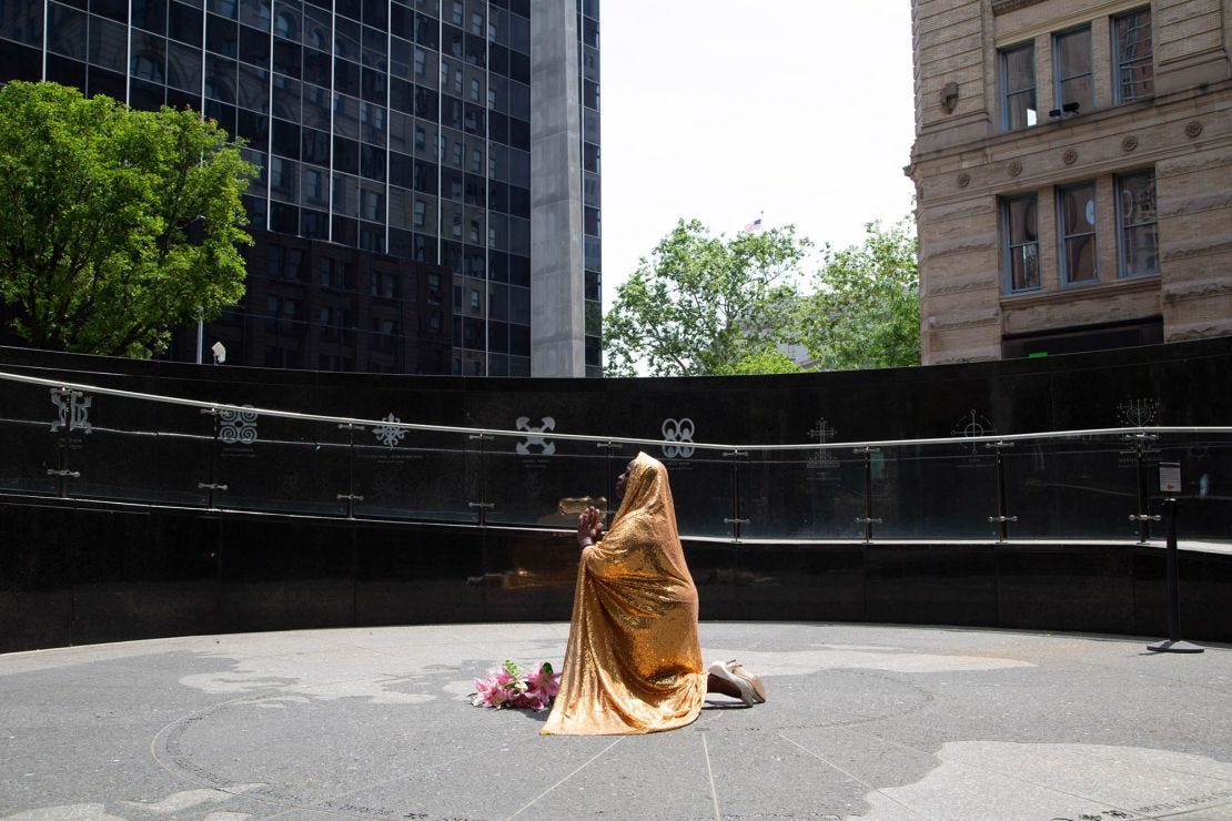 A golden cape Faustine wears in photos serves to represent the feminine divine, an energy that promotes positive change in the world and serves to protect sites that carry the legacies of the enslaved. Pictured above, Faustine poses at the African Burial Ground National Monument in downtown Manhattan. A golden cape Faustine wears in photos serves to represent the feminine divine, an energy that promotes positive change in the world and serves to protect sites that carry the legacies of the enslaved. Pictured above, Faustine poses at the African Burial Ground National Monument in downtown Manhattan.