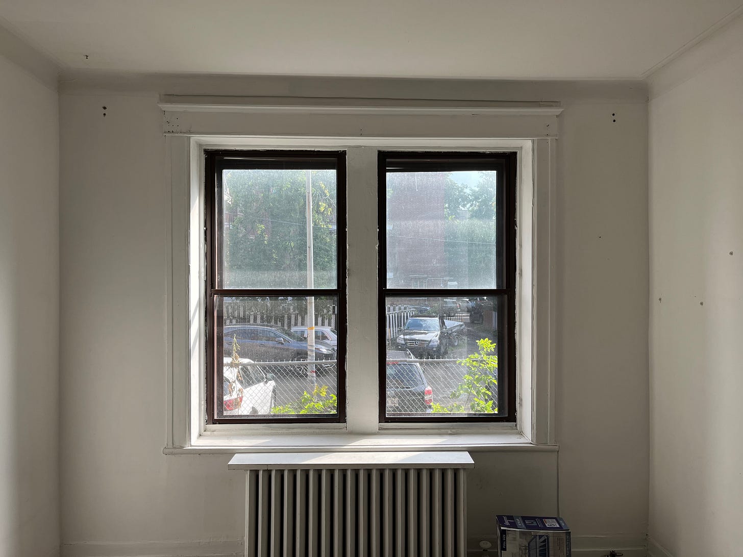 Two windows look out over a parking lot. They are above a radiator, in a very empty whitewashed room.