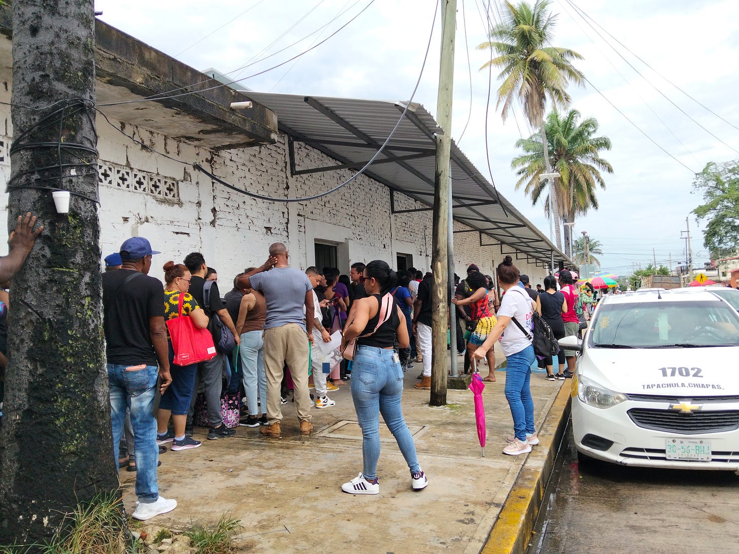 A line of people stand on the sidewalk by a white building