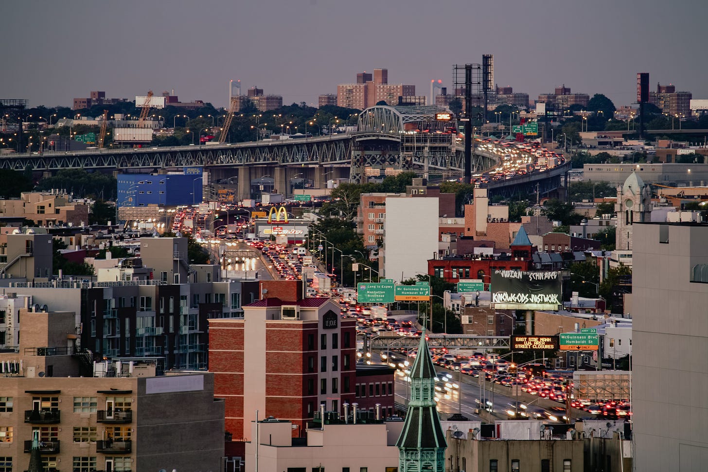 An elevated view over a dense city neighborhood at dusk. A major highway packed with cars cuts through the scene, with the red taillights of the traffic glowing brightly. A large bridge spans the background.