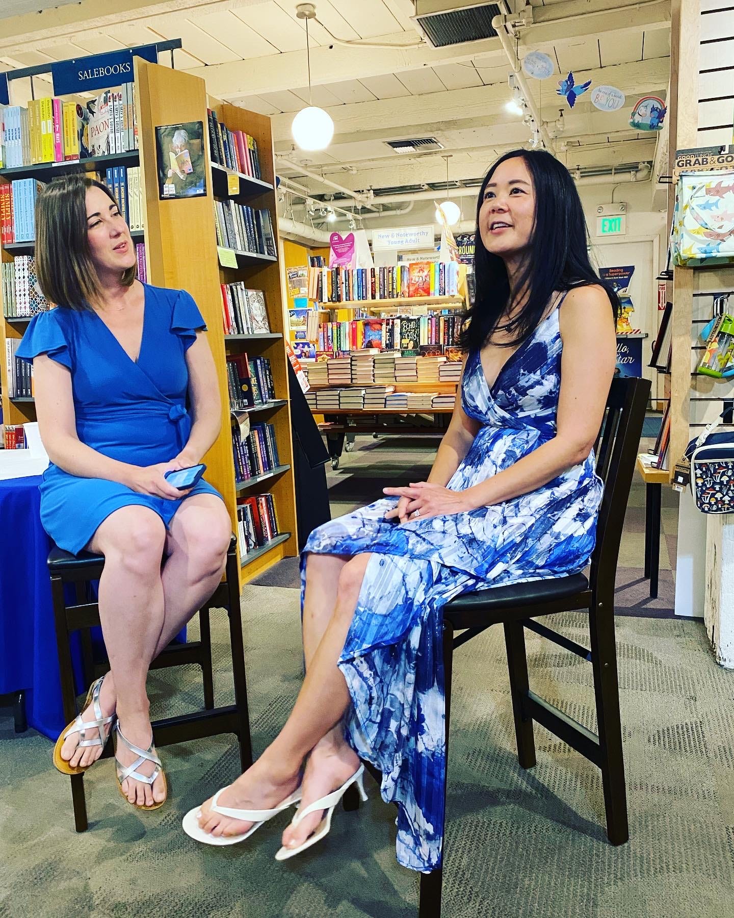 Joanna Phoenix in blue dress, interviewing Evelyn Skye in blue and white dress in a bookstore Joanna Phoenix in blue dress, interviewing Evelyn Skye in blue and white dress in a bookstore
