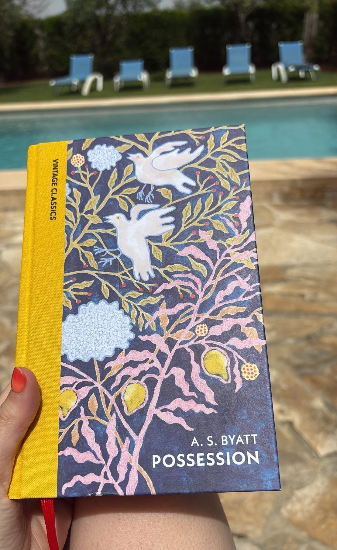 A woman's hand holds a hardback copy of Possession, which has a yellow fabric spine and an illustrated cover with doves landing in a tree with berries on the branches and the text: Possession, A.S. Byatt, Vintage Classics. In the background, there is a pool and a line of beach lounges across from where Caroline is reading.